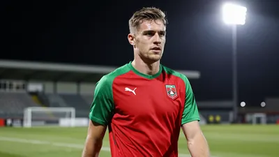 A determined, athletic 25-year-old FFK midfielder, with a strong build and focused expression, wears a plain red and...