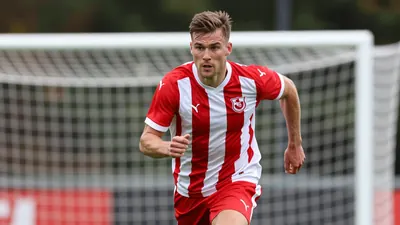 A Fredrikstad FK player in a red and white striped jersey, mid-20s, muscular build, sprinting towards the goal with...